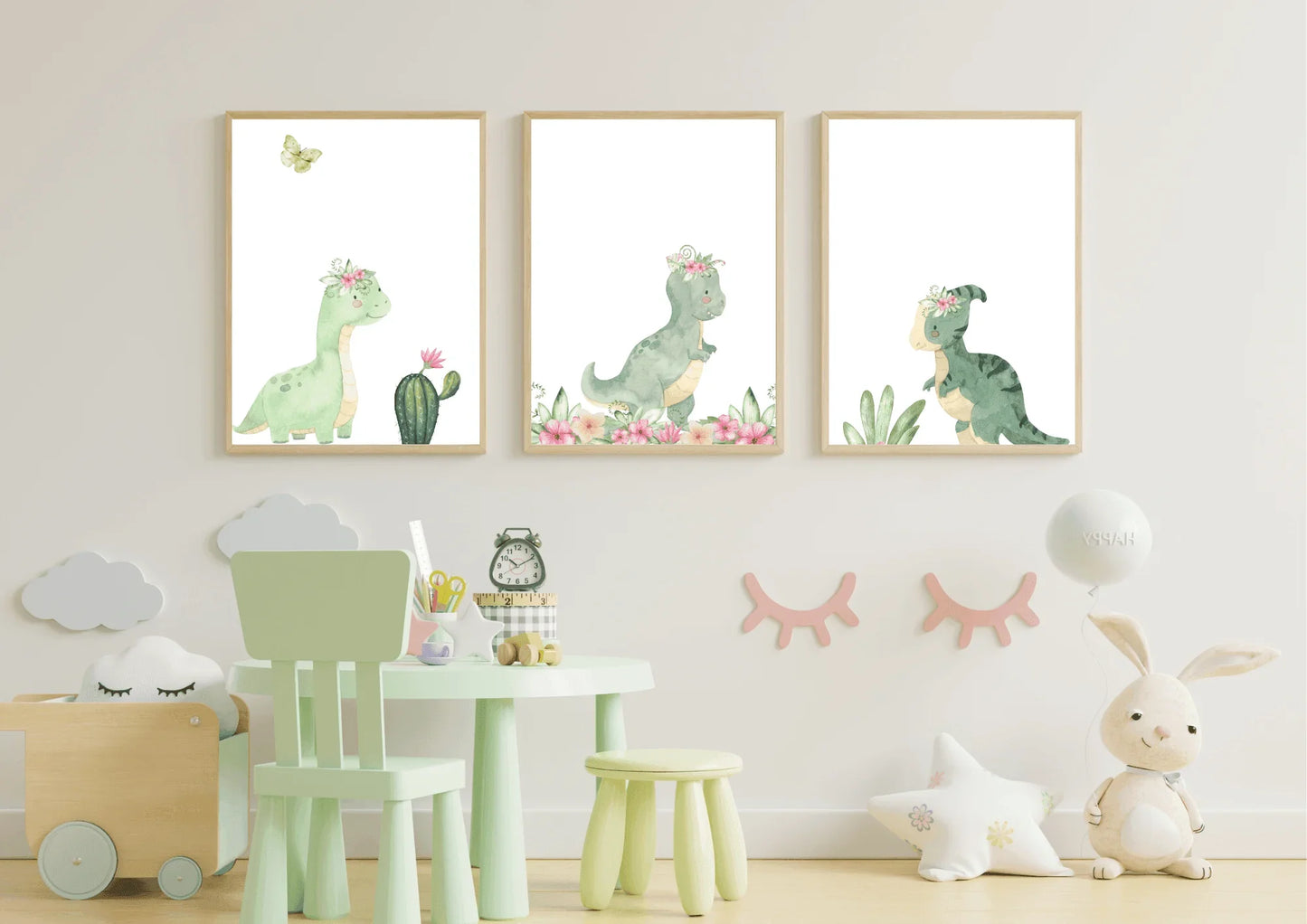 Three framed watercolor prints of cute green dinosaurs wearing pink floral crowns, displayed side-by-side on a white nursery wall. The prints feature a long-necked dinosaur, a T-Rex, and a smaller dinosaur. Below the artwork is a small, mint green child's table and chair set, a wooden toy box on the left, and a plush rabbit and star pillow on the right.