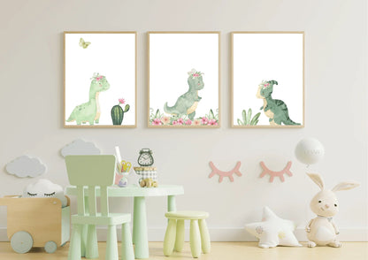 Three framed watercolor prints of cute green dinosaurs wearing pink floral crowns, displayed side-by-side on a white nursery wall. The prints feature a long-necked dinosaur, a T-Rex, and a smaller dinosaur. Below the artwork is a small, mint green child's table and chair set, a wooden toy box on the left, and a plush rabbit and star pillow on the right.