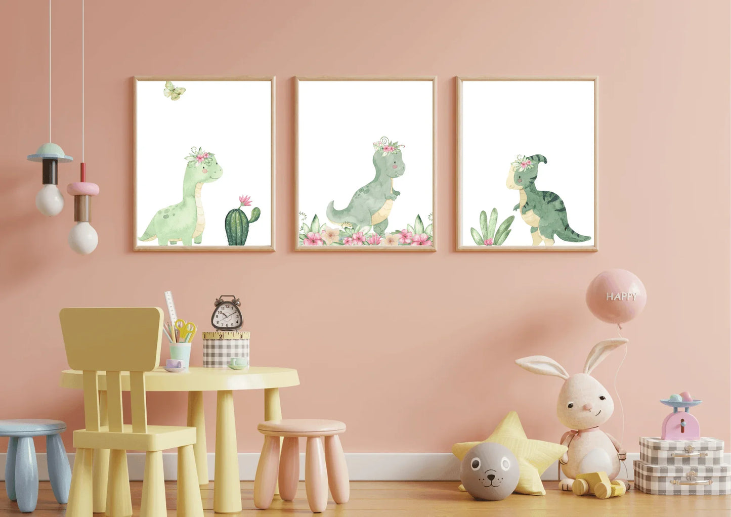 A set of three framed watercolor prints featuring cute green dinosaurs wearing pink floral crowns, displayed side-by-side on a light pink wall in a child's room. The prints include a long-necked dinosaur next to a cactus, a T-Rex with a flower border, and a smaller dinosaur with leafy plants. In the foreground is a small, pale yellow children's table with pink and blue stools, a yellow chair, and a plush bunny and toys on the floor to the right.