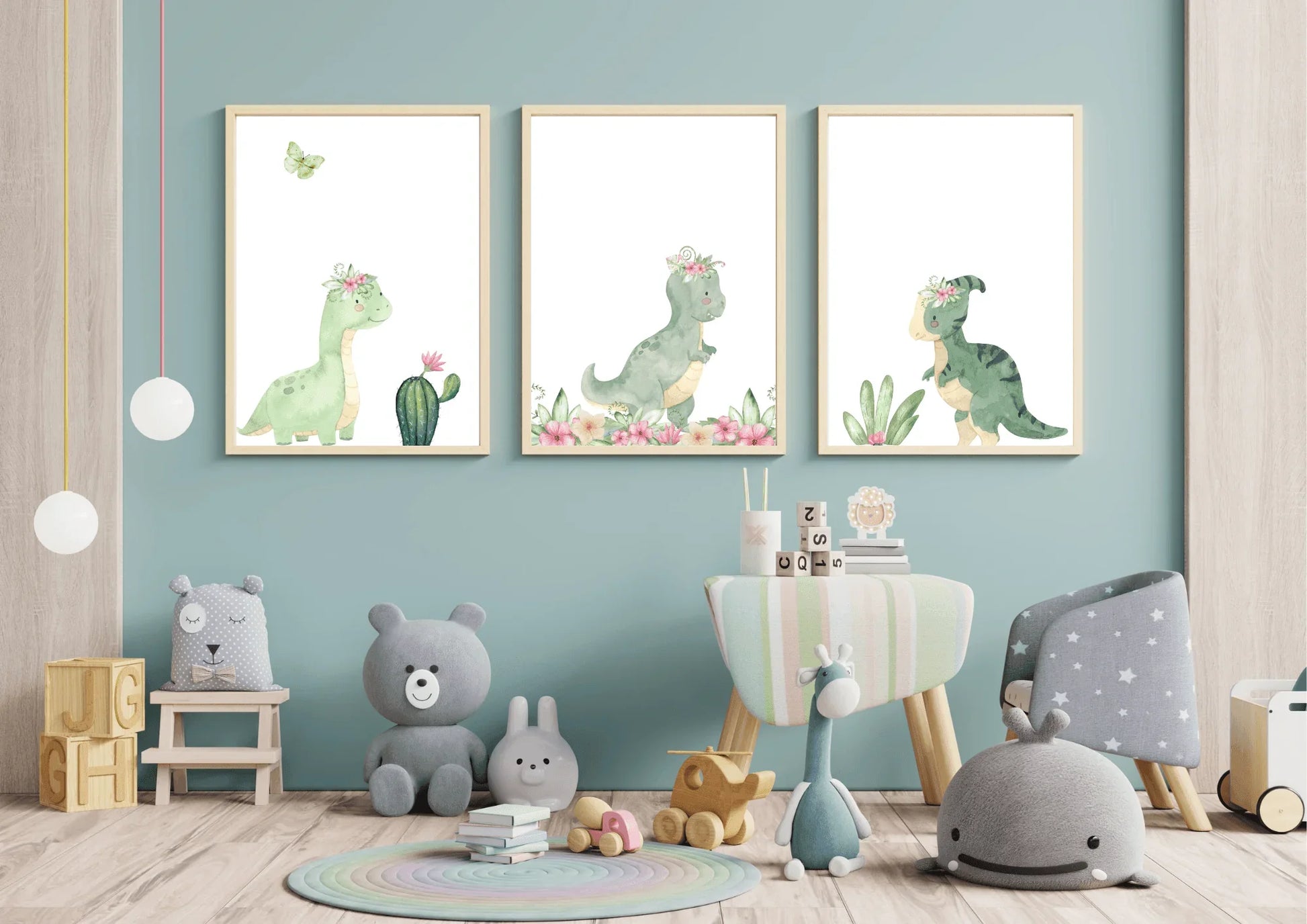 Three framed watercolor prints of cute green dinosaurs with pink floral crowns, hung side-by-side on a light teal-blue wall in a nursery. The prints feature a long-necked dinosaur, a T-Rex, and a smaller dinosaur. The floor is covered with a variety of soft toys, including gray stuffed animals, a blue giraffe, and a round rug with pastel stripes.