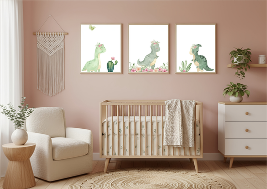 A set of three framed watercolor prints of cute green dinosaurs with floral crowns, displayed over a natural wood crib in a nursery with light dusty rose walls. To the left is a cream-colored armchair and a round wood side table with a plant. To the right is a white dresser with plants on a floating shelf above it. A macrame wall hanging is visible on the far left.