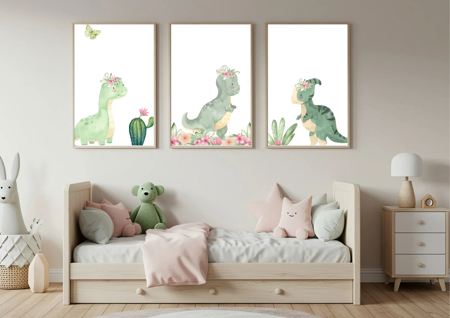 Three framed vertical watercolor prints of cute green dinosaurs wearing pink floral crowns, hung above a light wood toddler bed. The prints are against a neutral beige wall. The bed is dressed with a pale pink blanket and several plush pillows, including a stuffed green dinosaur. To the left is a large plush rabbit in a basket, and to the right is a small wooden nightstand with a lamp.