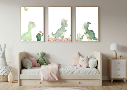 Three framed vertical watercolor prints of cute green dinosaurs wearing pink floral crowns, hung above a light wood toddler bed. The prints are against a neutral beige wall. The bed is dressed with a pale pink blanket and several plush pillows, including a stuffed green dinosaur. To the left is a large plush rabbit in a basket, and to the right is a small wooden nightstand with a lamp.