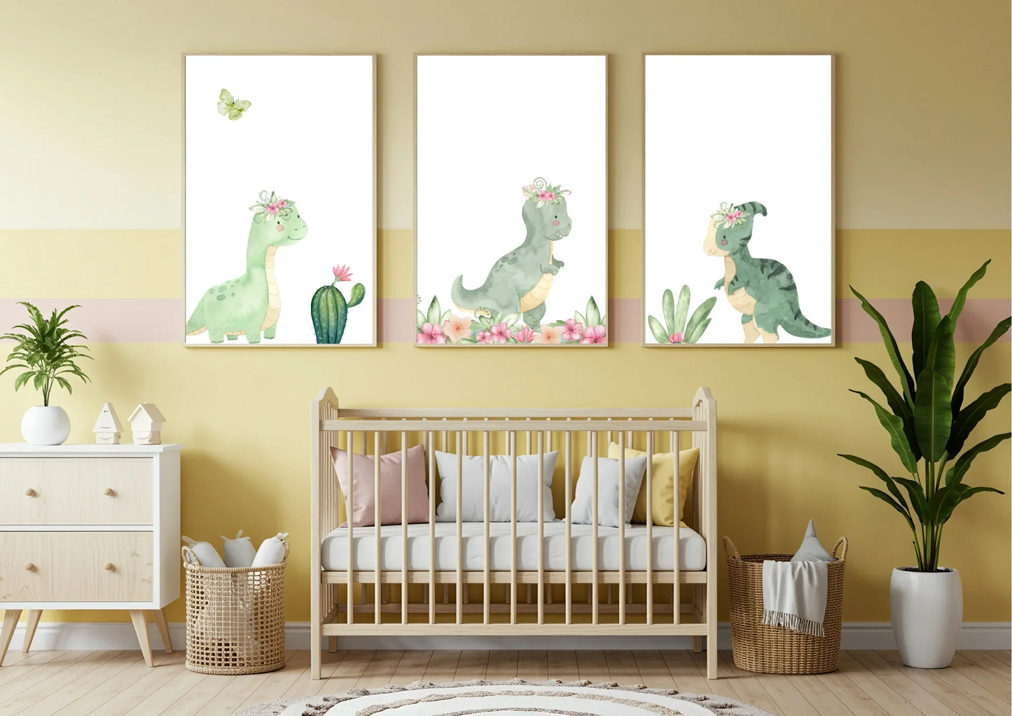 A set of three framed watercolor prints of cute green dinosaurs with pink floral crowns, hung on a yellow wall above a light wood crib. The crib is decorated with pink, white, and yellow pillows. To the left is a white and wood dresser with a plant and woven baskets, and to the right is a tall green houseplant in a white pot, next to a matching woven basket.
