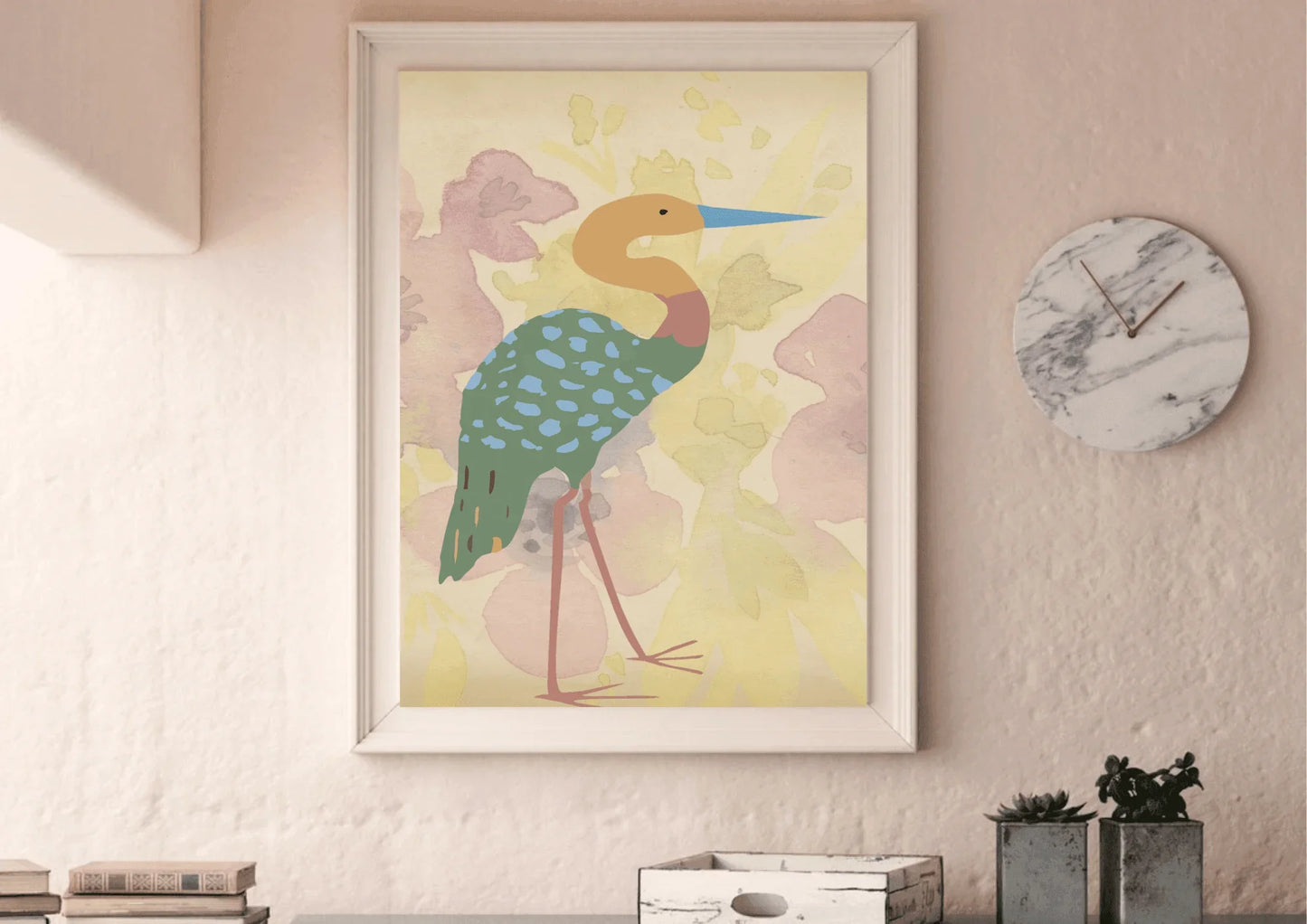 A framed digital art print of a stylized bird, which appears to be a heron, with a long blue beak, an orange neck, and a turquoise body with light blue spots. The background is a soft watercolor wash of pinks and yellows. The print is hanging on a light pink textured wall above a rustic wooden desk with a marble clock and small plants.
