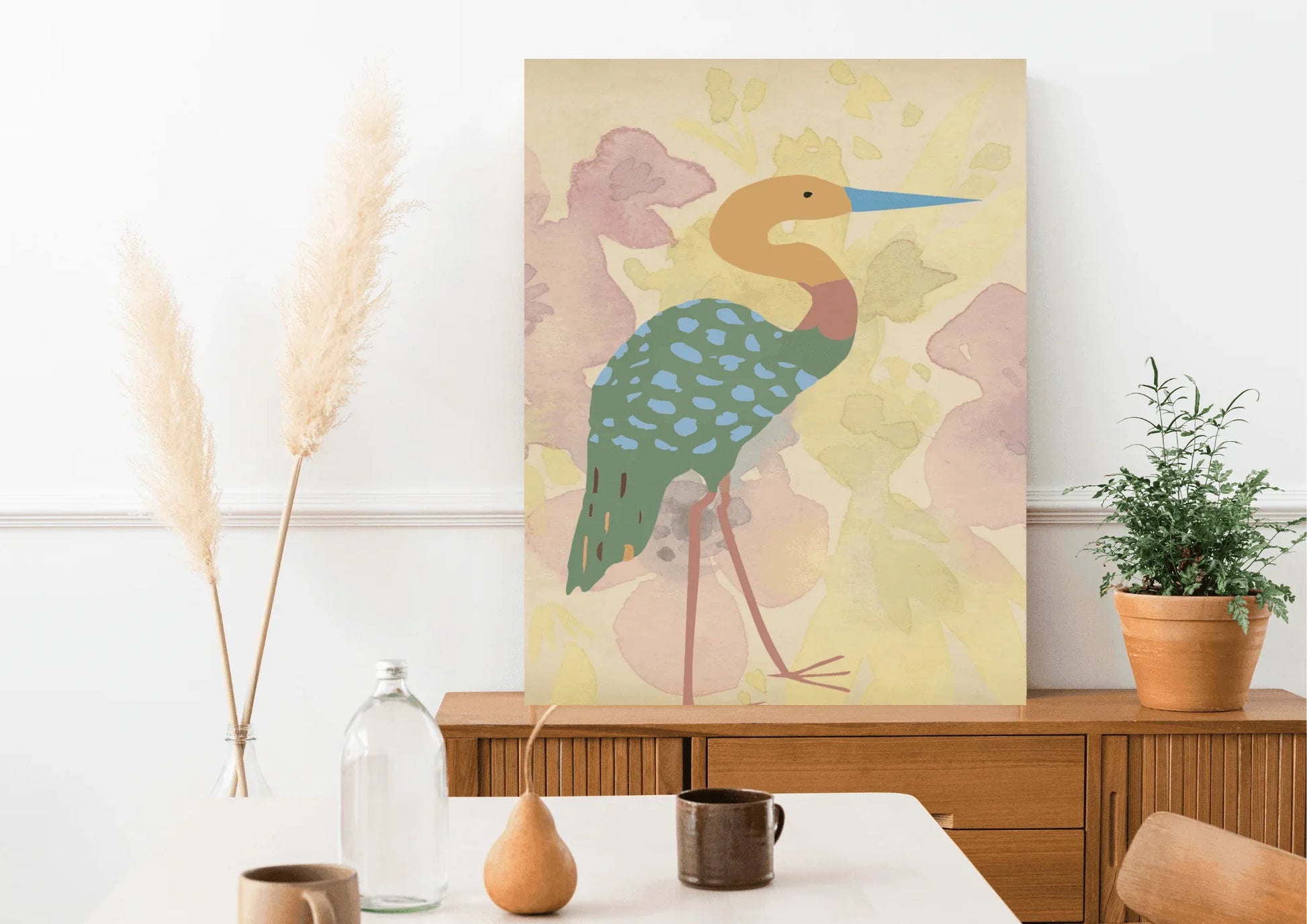 A framed digital art print of a stylized heron, with a blue beak and a turquoise spotted body, leaning against a wooden mid-century modern credenza. A vase with pampas grass is on the left, and a potted plant is on the right.