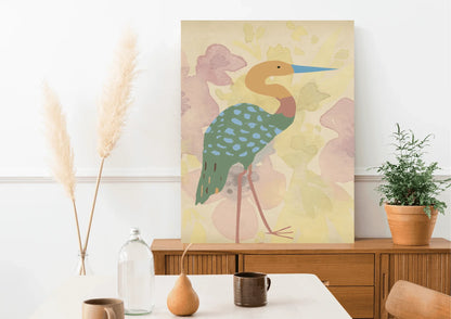 A framed digital art print of a stylized heron, with a blue beak and a turquoise spotted body, leaning against a wooden mid-century modern credenza. A vase with pampas grass is on the left, and a potted plant is on the right.