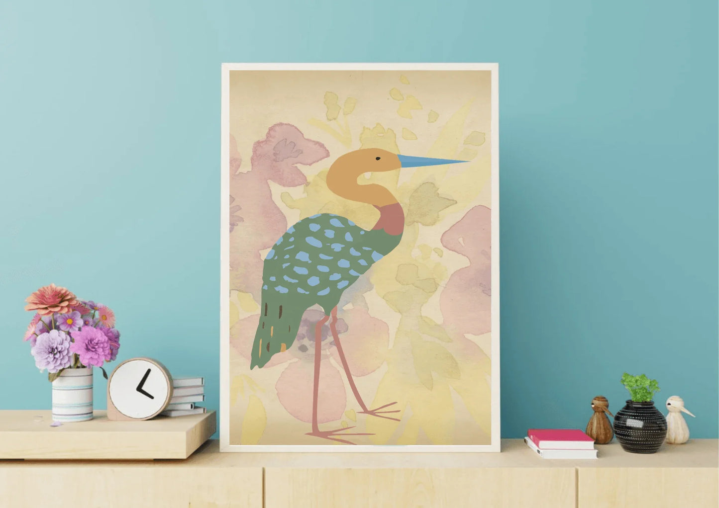 A framed digital art print of a stylized heron, with a blue beak and a turquoise spotted body, leaning against a light wood cabinet. The cabinet has a vase of flowers, a small clock, and books on top, all placed against a light blue wall.