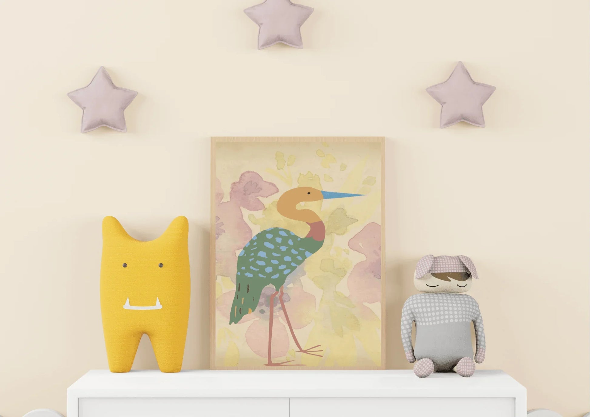 A framed digital art print of a stylized heron, with a blue beak and a turquoise spotted body, leaning against a white dresser in a child's room. A yellow plush toy and a gray and white stuffed rabbit sit on either side of the print. Three pale pink star decorations are on the wall above the dresser.