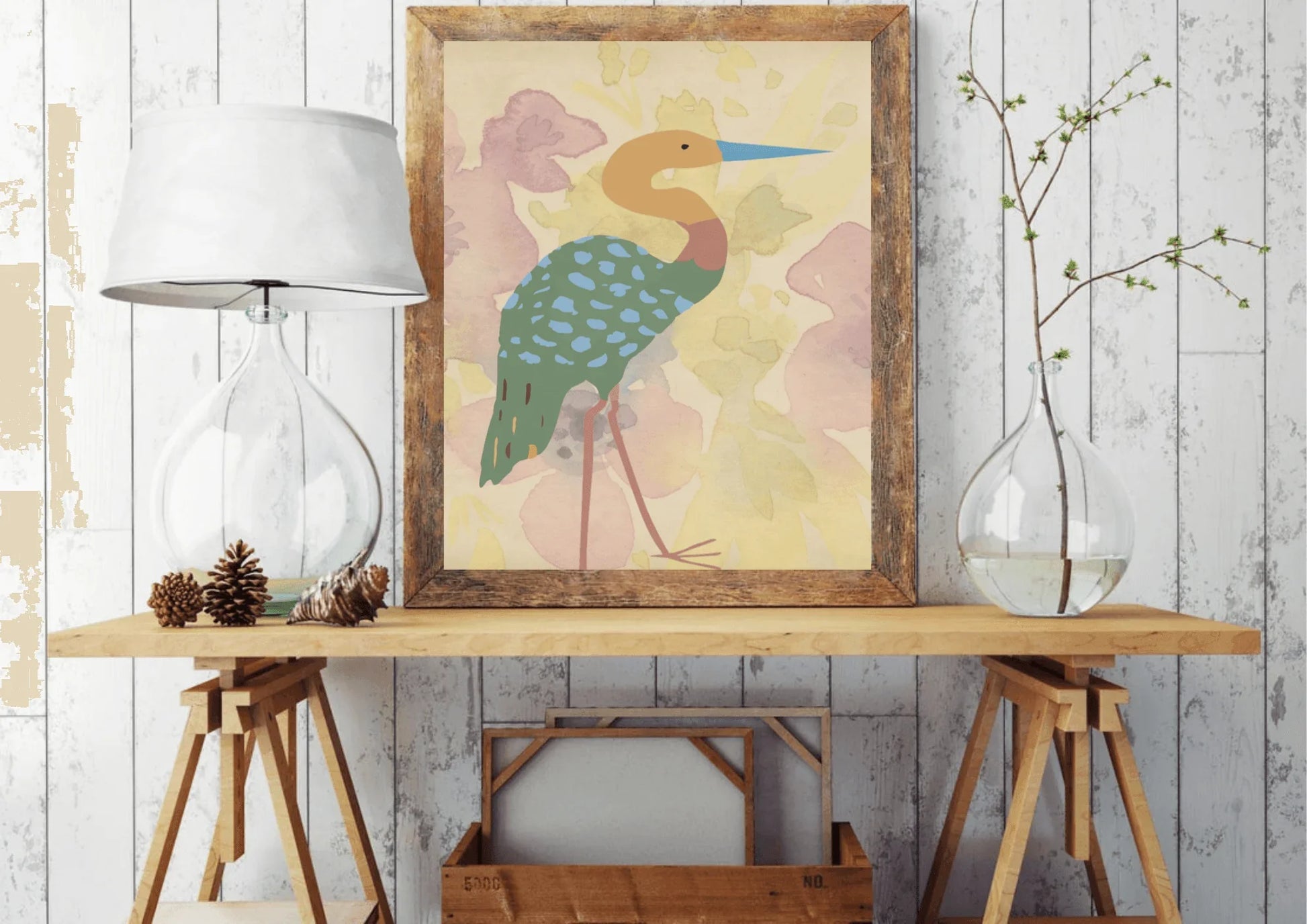 
A framed digital art print of a stylized heron, with a blue beak and a turquoise spotted body, leaning against a rustic white wooden plank wall. The print is on a raw wood table between a glass lamp and a clear vase with a tree branch.