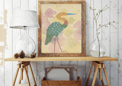 
A framed digital art print of a stylized heron, with a blue beak and a turquoise spotted body, leaning against a rustic white wooden plank wall. The print is on a raw wood table between a glass lamp and a clear vase with a tree branch.
