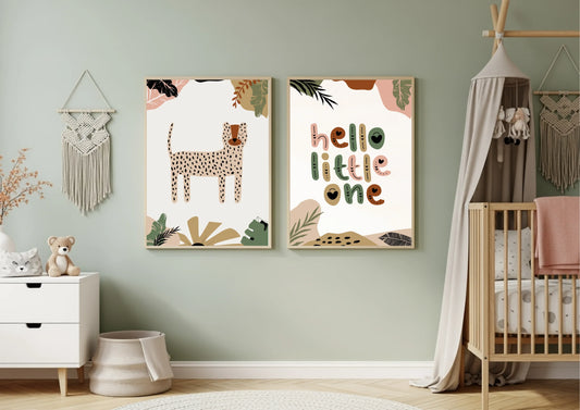 A nursery with pale green walls, featuring a two-piece framed art set with a boho jungle theme. The prints include a spotted cheetah/leopard and the phrase "hello little one" in earthy tones. The room has a white dresser, a macrame wall hanging, and a wooden crib with a canopy.
