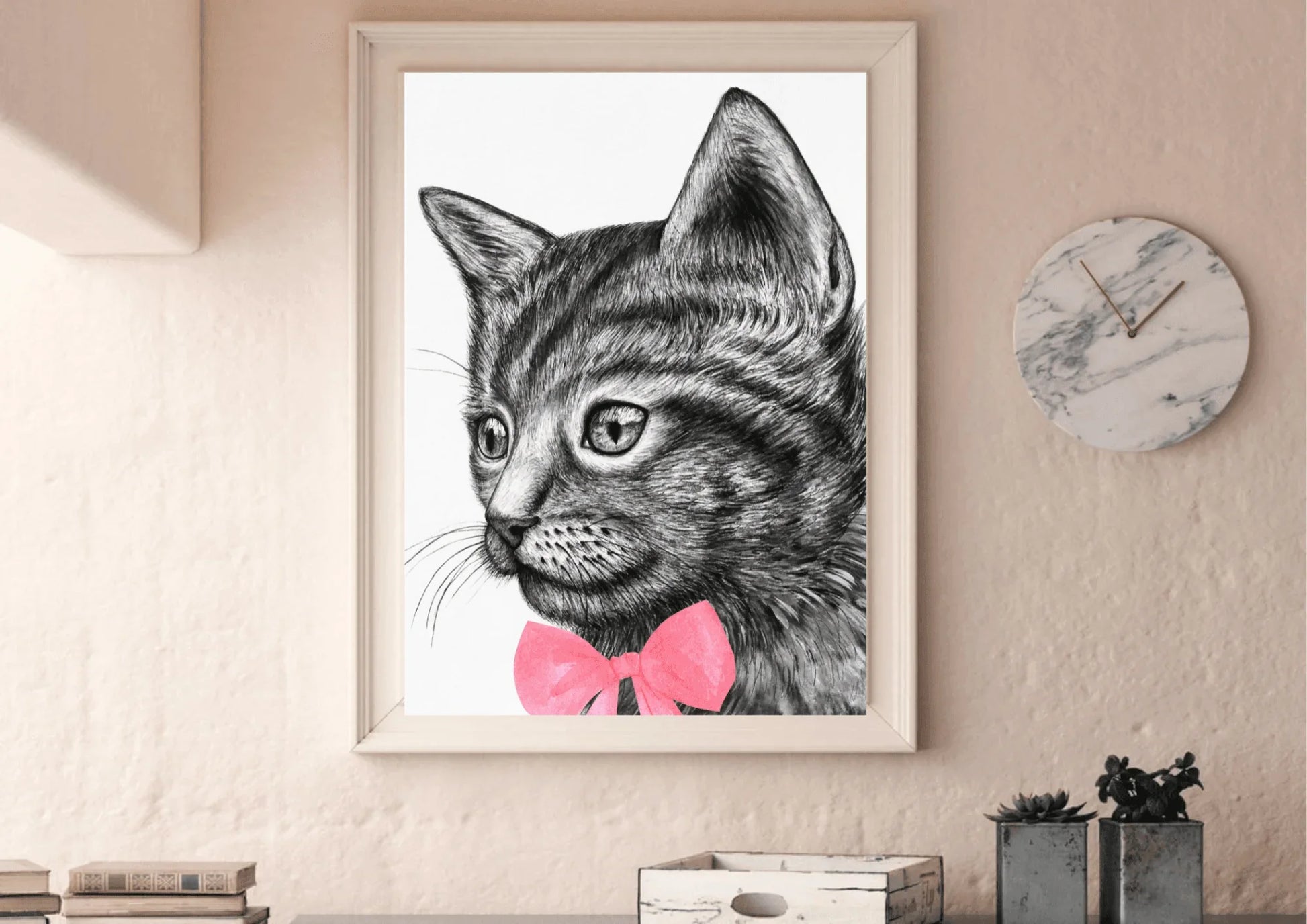 
A framed digital art print of a black and white cat with a pink bow tie hanging on a textured, light-colored wall above a wooden desk. A round marble clock and small potted succulents are visible on the right.