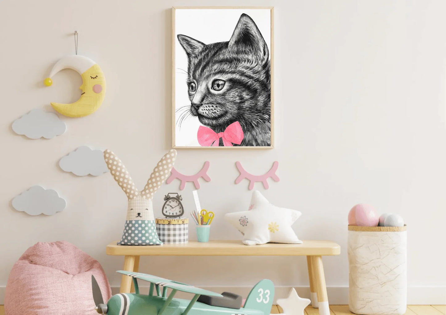 A framed digital art print of a black and white cat with a pink bow tie hanging on a white wall in a child's room. The room has a small wooden table with a plush bunny, a star-shaped pillow, and a toy airplane on the floor.