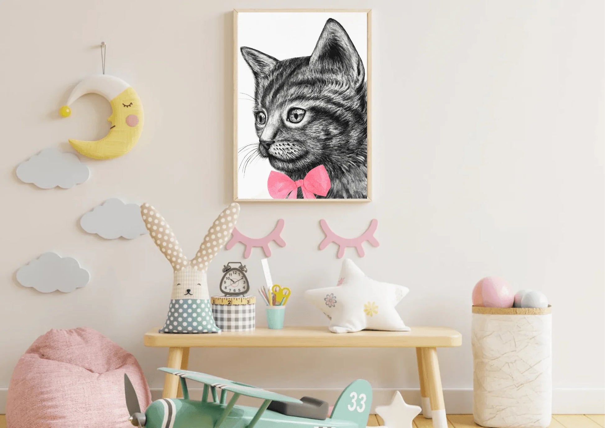 A framed digital art print of a black and white cat with a pink bow tie hanging on a white wall in a child's room. The room has a small wooden table with a plush bunny, a star-shaped pillow, and a toy airplane on the floor.