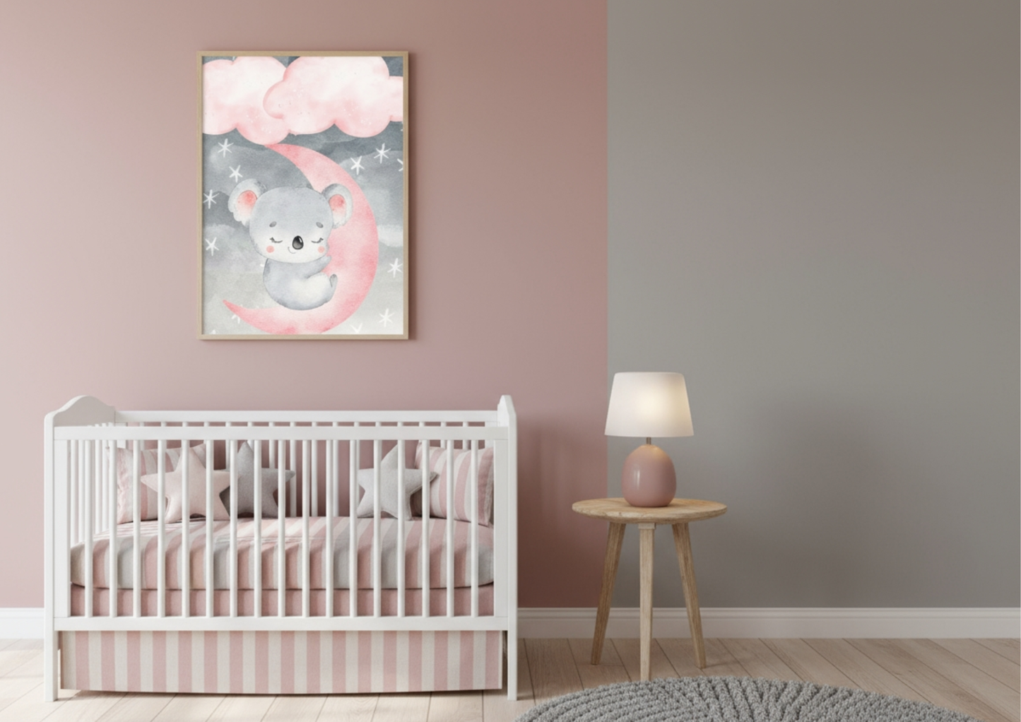 A framed vertical watercolor print featuring a sweet sleeping koala resting on a bright pink crescent moon, surrounded by grey clouds and tiny stars. The print hangs on a wall that is painted in two sections: a dusky rose pink on the left and a neutral taupe grey on the right. Below the artwork is a white wooden crib dressed with striped pink and white bedding. To the right of the crib is a round wooden side table holding a soft pink lamp. The room is styled with a plush grey circular rug and is focused on 
