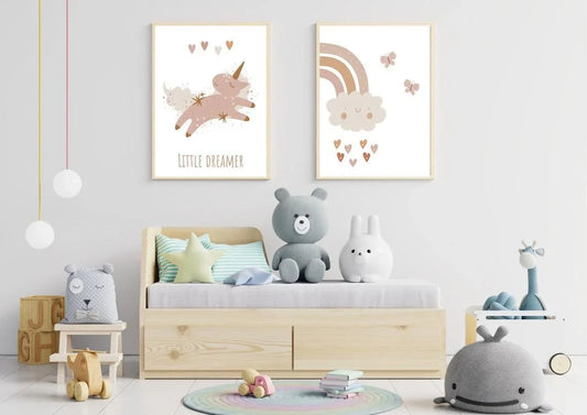unicor
A close-up of two framed art prints hanging above a child's bed. The first print features a whimsical unicorn and the text "LITTLE DREAMER" in a muted, boho color palette. The second print depicts a smiling cloud with a rainbow and falling hearts. A large gray teddy bear and a white rabbit toy sit on the bed. nursery print