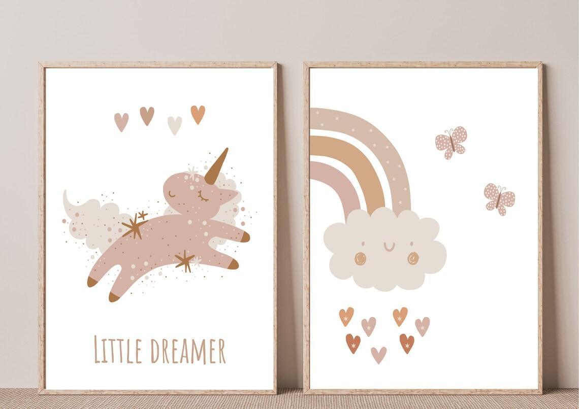 
A close-up of two framed art prints leaning against a beige wall. The first print features a whimsical unicorn with a "LITTLE DREAMER" text in a muted, boho color palette. The second print shows a smiling cloud with a rainbow and hearts in a similar style.