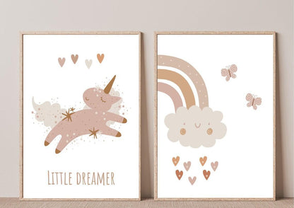 
A close-up of two framed art prints leaning against a beige wall. The first print features a whimsical unicorn with a "LITTLE DREAMER" text in a muted, boho color palette. The second print shows a smiling cloud with a rainbow and hearts in a similar style.