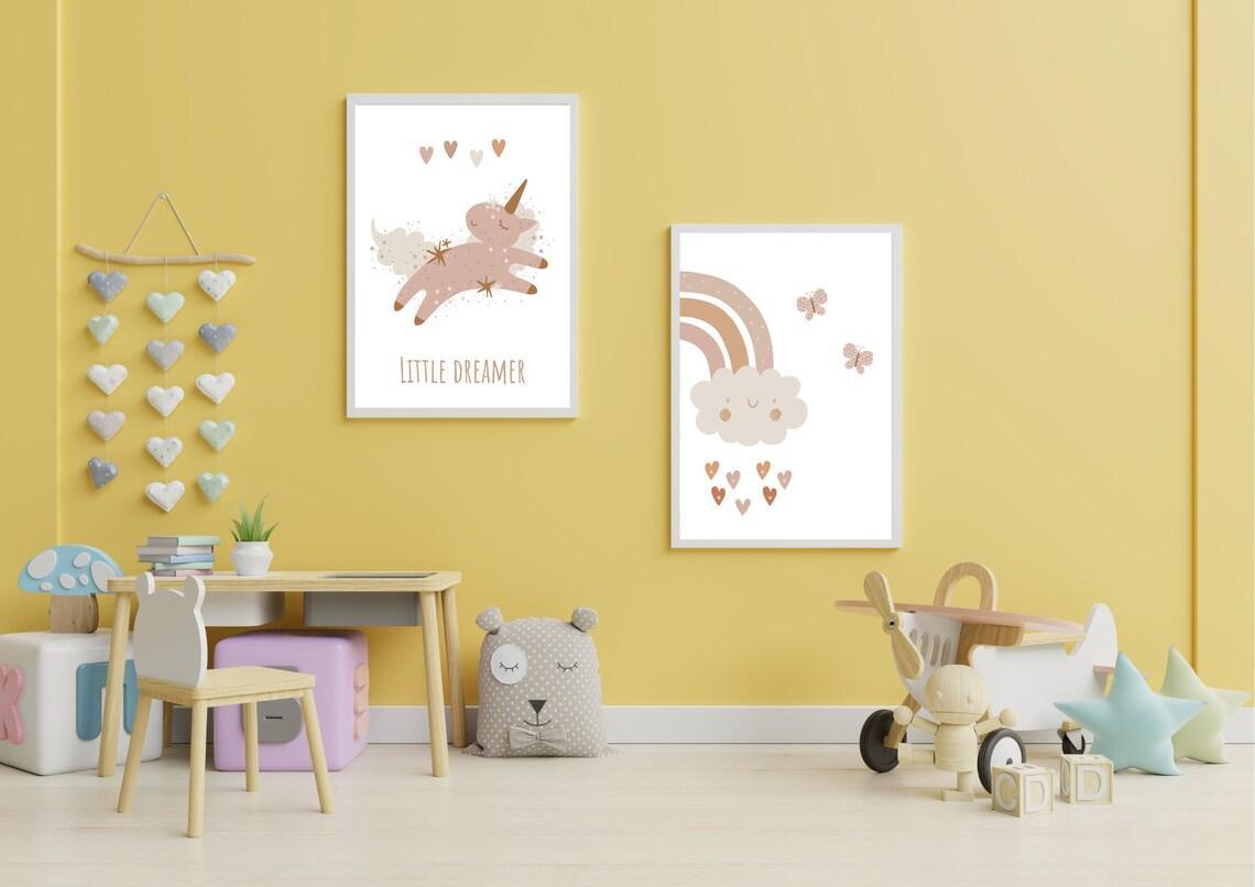 
A set of two framed art prints hangs on a bright yellow wall in a child's playroom. The first print features a whimsical unicorn and the text "LITTLE DREAMER" in a muted, boho color palette. The second print depicts a smiling cloud with a rainbow and falling hearts. The room is decorated with a small table and chair set, a rocking plane toy, and heart-shaped wall hangings.
