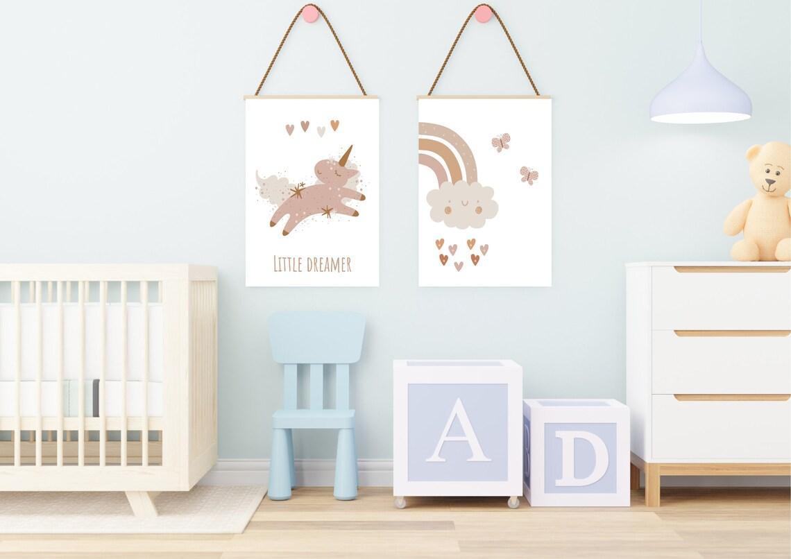 A set of two art prints, hanging from ropes with wooden rods, on a light blue wall in a baby's room. The print on the left shows a whimsical unicorn with the phrase "LITTLE DREAMER" and the print on the right depicts a smiling cloud with a rainbow and hearts. A wooden crib, a small blue chair, and large letter blocks are visible below the prints.
