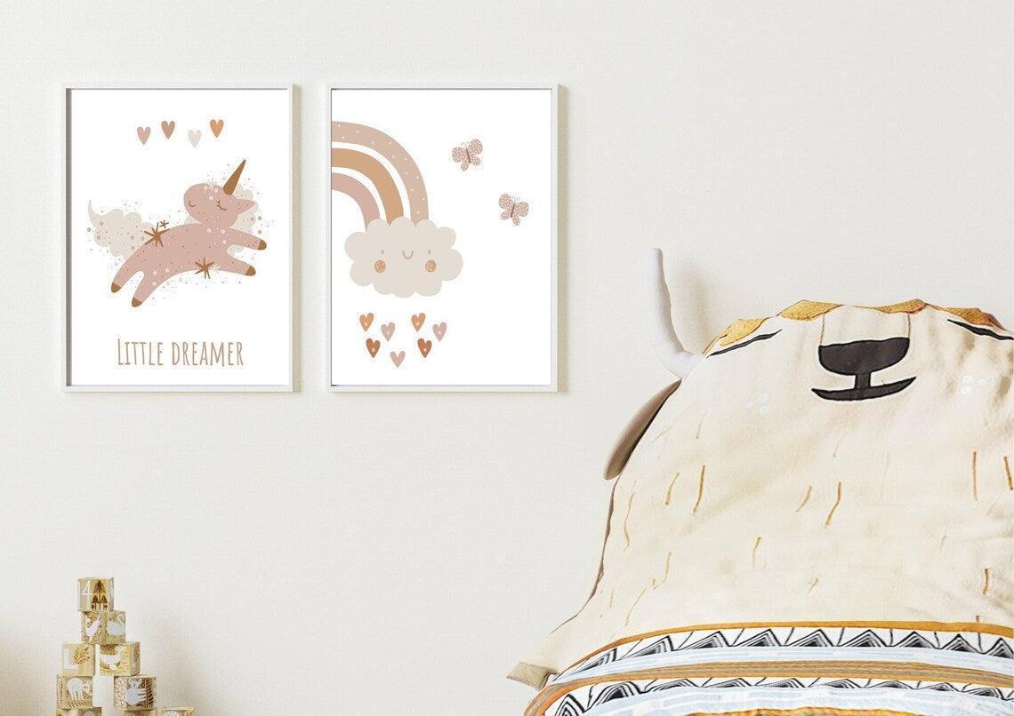
A set of two framed art prints hangs on a white wall above a child's bed. The first print features a whimsical unicorn with the text "LITTLE DREAMER" and the second print depicts a smiling cloud with a rainbow and hearts. The corner of a pillow with a bull or cow face design is visible on the right side of the image.
