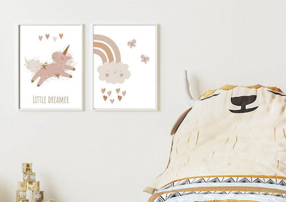 
A set of two framed art prints hangs on a white wall above a child's bed. The first print features a whimsical unicorn with the text "LITTLE DREAMER" and the second print depicts a smiling cloud with a rainbow and hearts. The corner of a pillow with a bull or cow face design is visible on the right side of the image.