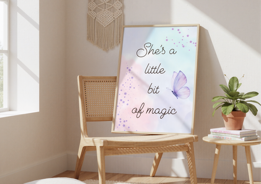 A sunny, modern boho bedroom with a light-colored wooden floor. A framed art print with a watercolor background and the words "She's a little bit of magic" is resting against a soft pink wall. The artwork is leaning on a simple, light-colored wooden chair. In the background, a large window with sheer white curtains allows sunlight to fill the room, and a string of pastel-colored pom-poms hangs down the side of the curtains.