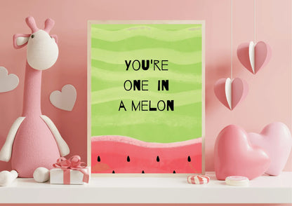 A framed "YOU'RE ONE IN A MELON" print with a watermelon design, identical to the first. It is set against a pink background on a white shelf. The print is surrounded by Valentine's Day-themed decor, including a tall, soft pink stuffed giraffe, a small pink gift box tied with a bow, hanging pink paper hearts, and several large pink heart decorations on the shelf.