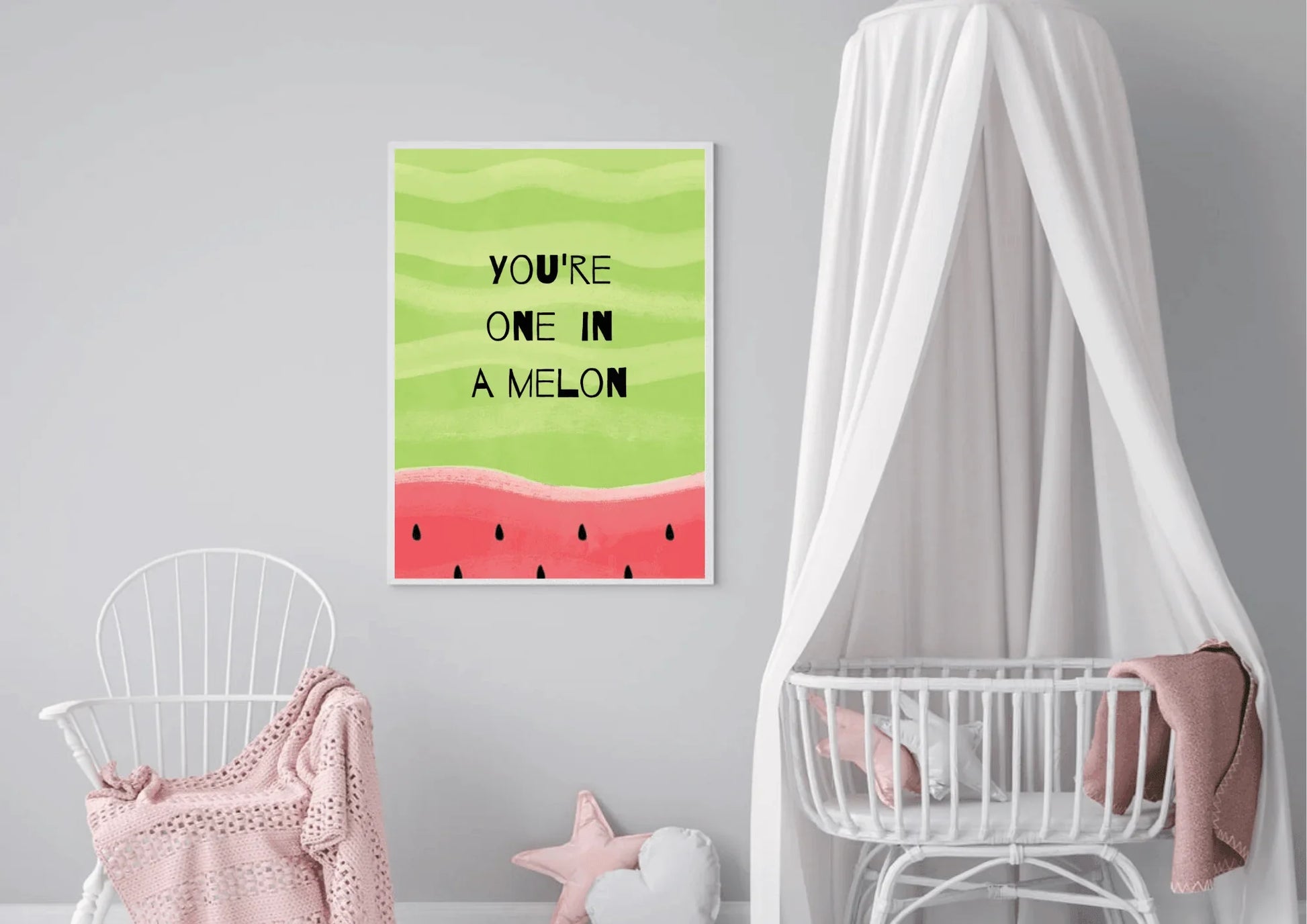 A framed "YOU'RE ONE IN A MELON" print with a watermelon design, centered on a light gray wall in a nursery or child's room. To the left is a white wicker rocking chair draped with a soft pink blanket. To the right is a white crib with a sheer white canopy draped over it. A pink star-shaped pillow rests on the floor.