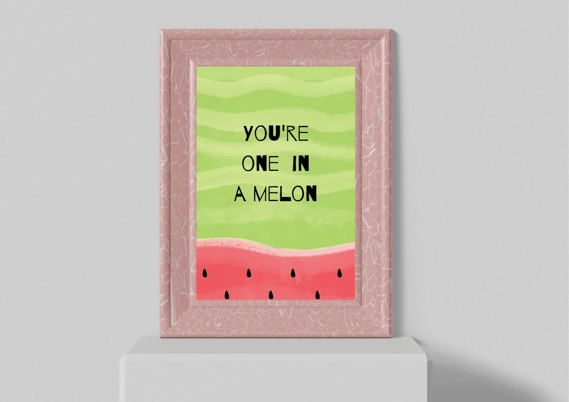 A close-up of the "YOU'RE ONE IN A MELON" watermelon print, displayed in a light pink frame with a distressed or cracked marble texture. The framed print is resting on a small, white, flat shelf against a plain gray wall.