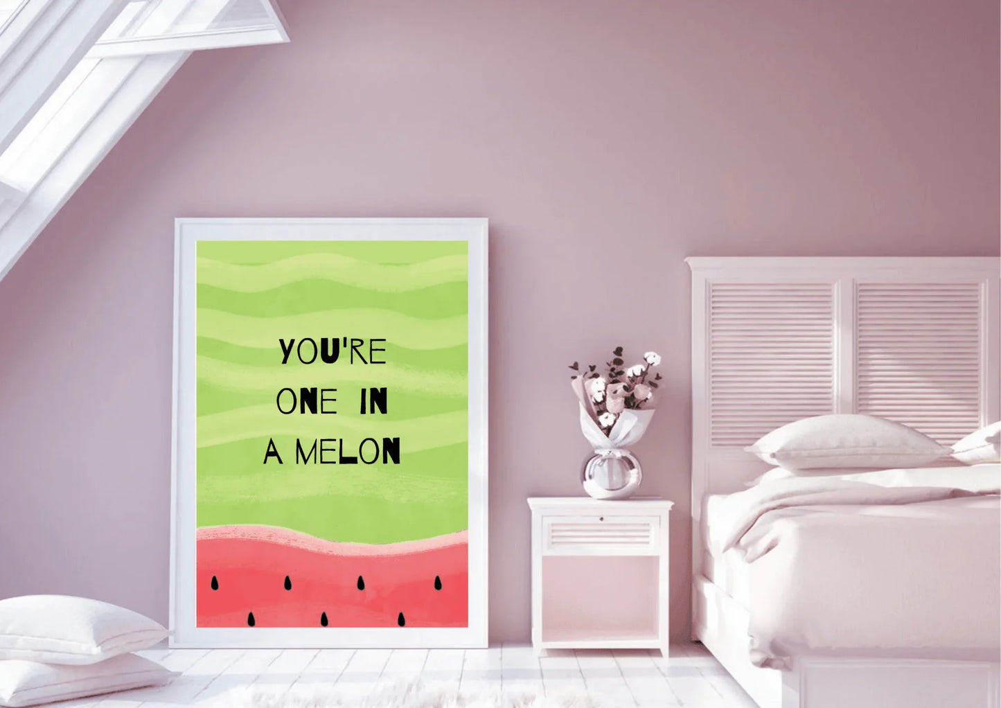A framed "YOU'RE ONE IN A MELON" watermelon print displayed in a white frame on the floor of a bedroom. The room features soft pink walls and a sloped ceiling with a window. To the right is a bed with a white headboard and pink bedding, next to a small white nightstand holding a clear vase with flowers. The room has soft lighting and a light wooden floor.