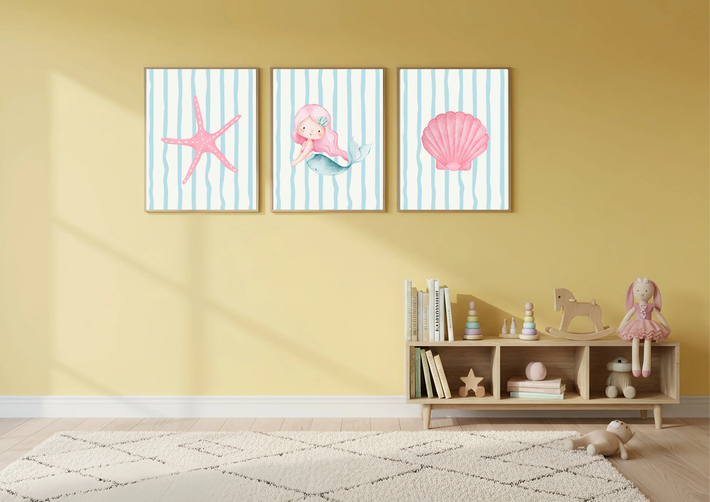 Three framed artworks with a starfish, seashell, and mermaid on a yellow wall above a wooden shelf with toys.
