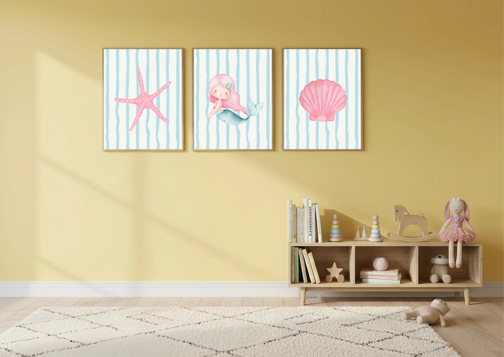 Three framed artworks with a starfish, seashell, and mermaid on a yellow wall above a wooden shelf with toys.
