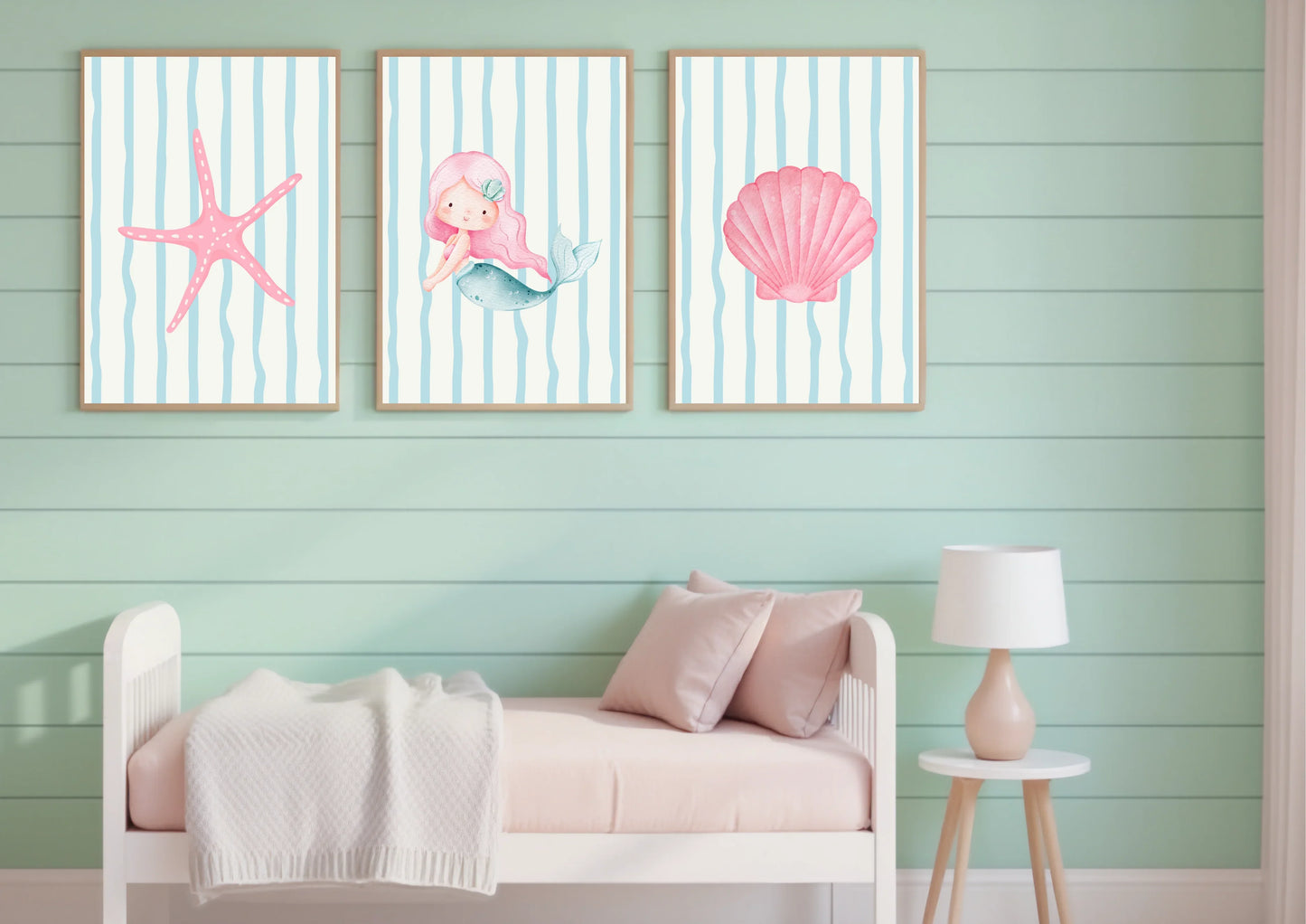 Three framed artworks of a mermaid, starfish, and shell on a mint green wall above a bed.