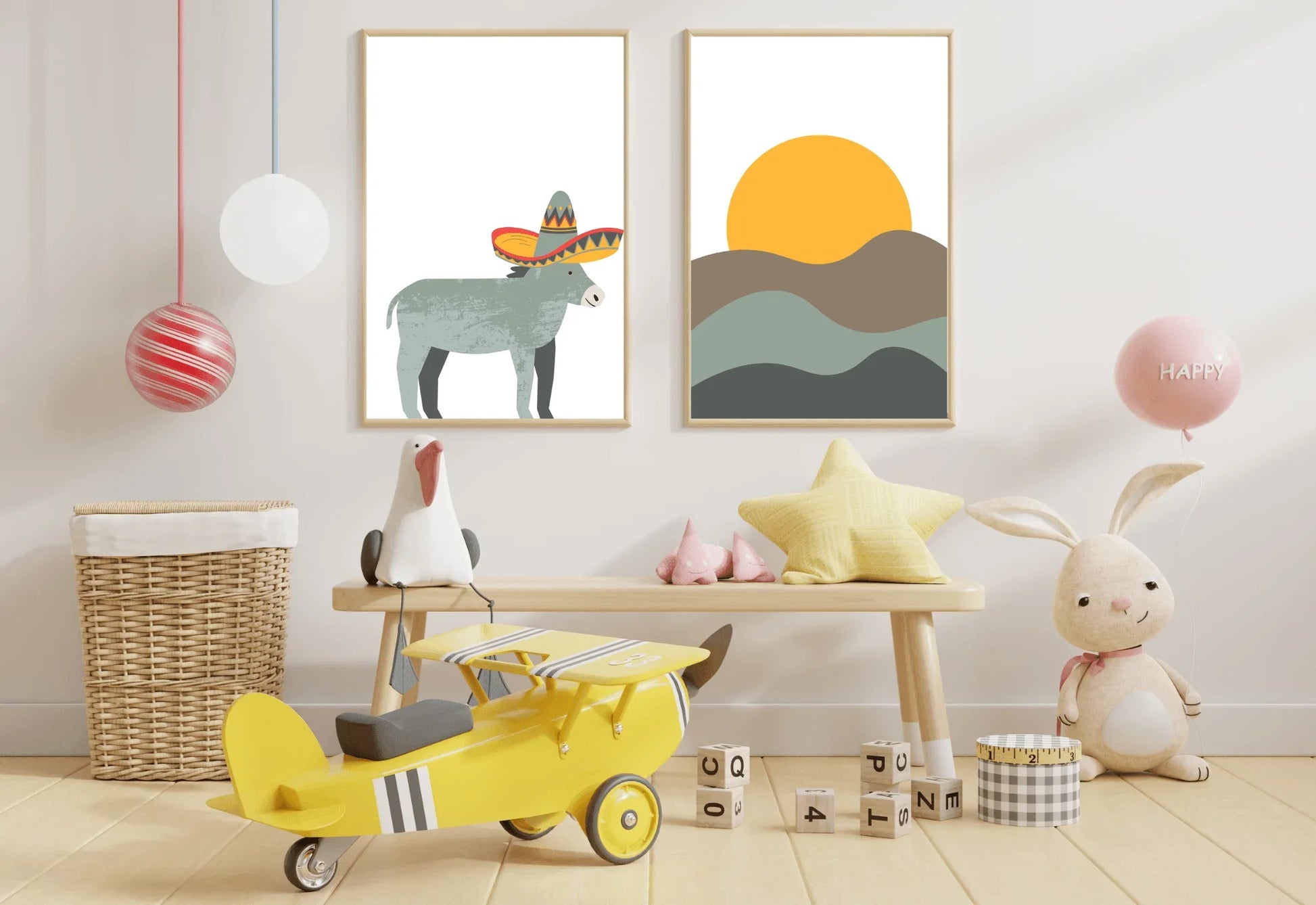 A colorful child's room setup featuring two framed art prints on a white wall: one of a gray donkey wearing a sombrero, and one of an orange sun over rolling brown and gray hills. The prints are surrounded by toys, including a large yellow toy airplane in the foreground, a light wood bench holding plush toys (a seagull and a yellow star), a wicker basket, a stuffed rabbit, and a pink "HAPPY" balloon.