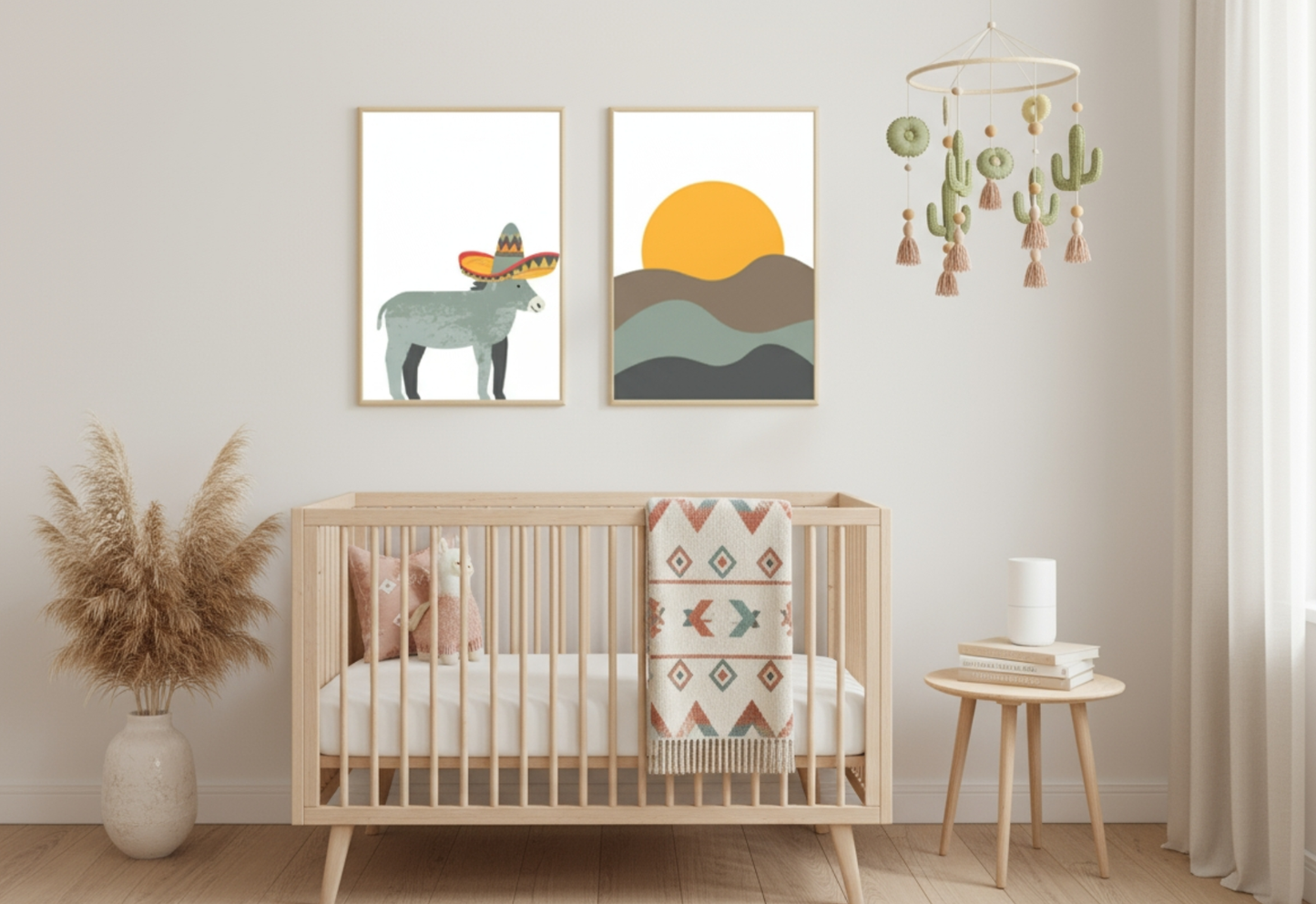 A Boho nursery with light-colored walls and wood flooring. Two framed prints hang above a light wood crib: one of a donkey wearing a sombrero, and one of a sunset over stylized desert hills. The crib has a blanket with a Southwestern pattern draped over the railing. To the left is a white vase with pampas grass, and to the right, a small side table holds books and a white candle. A cute cactus-themed mobile hangs above the crib.