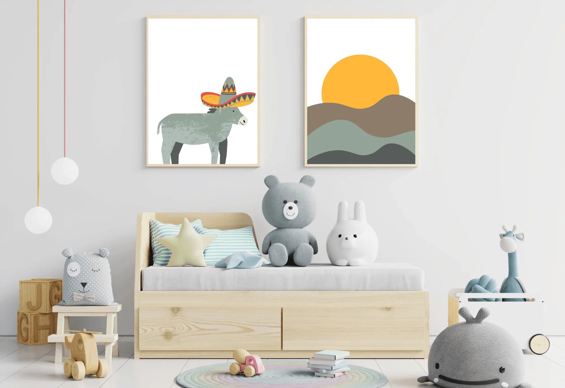 A child's room featuring a low light-wood bed with drawers underneath, decorated with several stuffed animals. Above the bed, two framed prints hang side-by-side: one of a gray donkey wearing a sombrero, and one of an orange sun over rolling brown and teal-green hills. The foreground includes various toys, such as a wooden cart, stacking blocks, and pendant lights hanging from the ceiling.