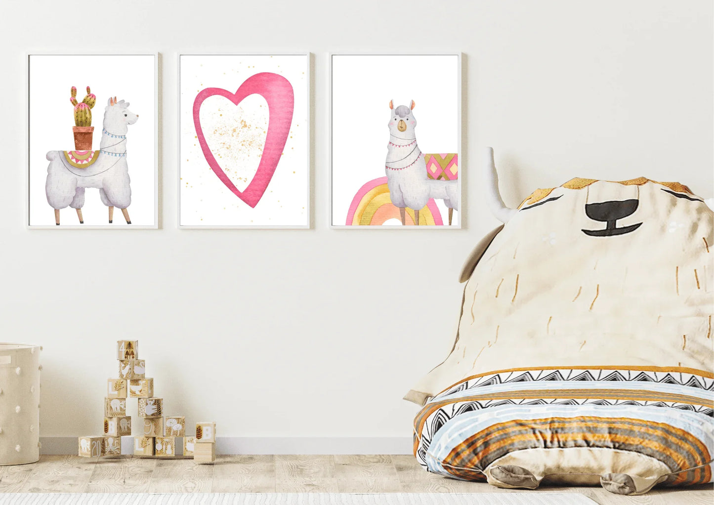 A trio of framed digital art prints featuring two llamas and a heart, hanging on a white wall in a child's room. A large, plush llama-shaped beanbag chair and a stack of wooden blocks are on the light wood floor.