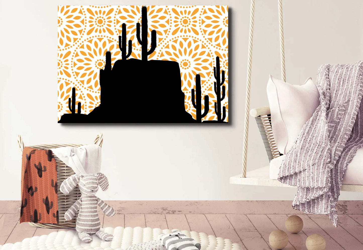 
A canvas art print with a black silhouette of a desert landscape featuring saguaro cacti and a rocky mesa. The background is a bright orange mandala pattern with a white circular design. The print hangs on a white wall above a large wicker basket and a small hanging swing chair.