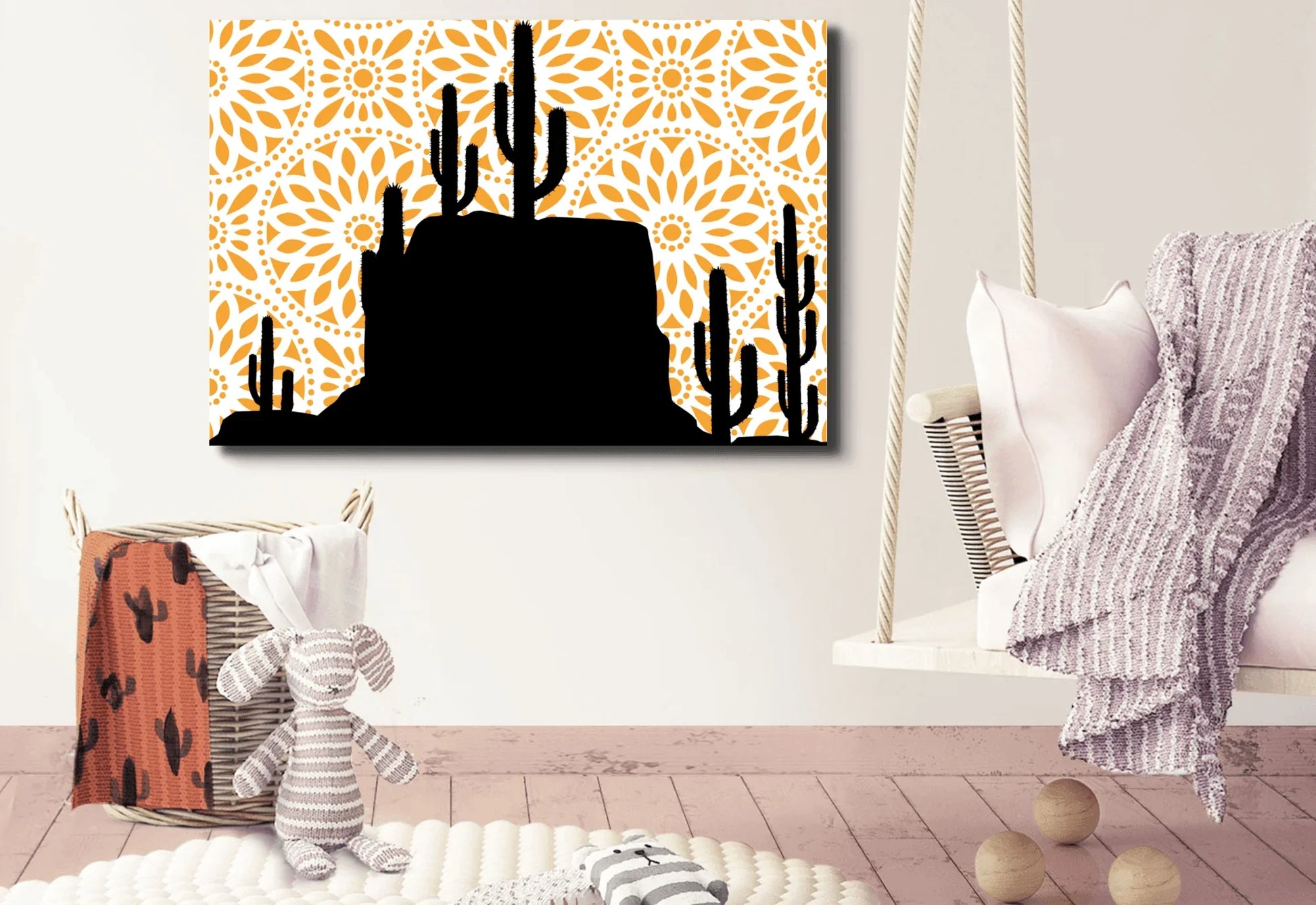 
A canvas art print with a black silhouette of a desert landscape featuring saguaro cacti and a rocky mesa. The background is a bright orange mandala pattern with a white circular design. The print hangs on a white wall above a large wicker basket and a small hanging swing chair.