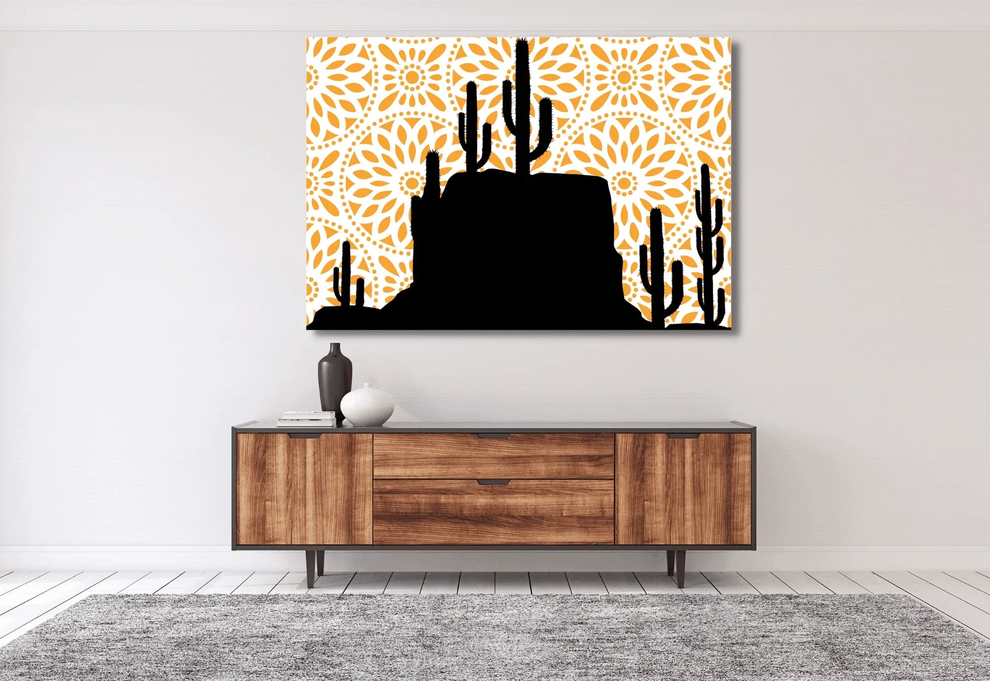 A large canvas art print featuring a black silhouette of a desert landscape with saguaro cacti and a rocky mesa. The background is a bright orange mandala pattern with a white circular design. The print hangs on a white wall above a long wooden mid-century modern credenza.