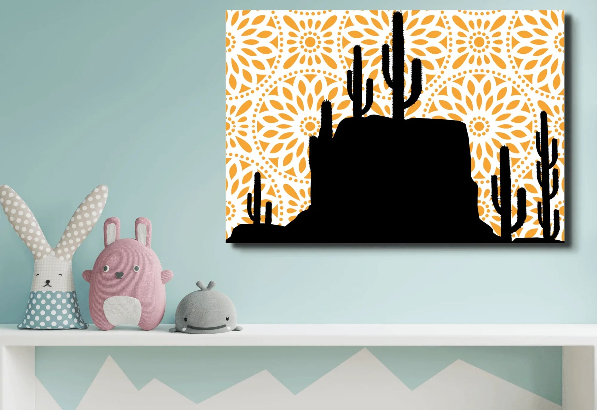 A large canvas art print featuring a black silhouette of a desert landscape with saguaro cacti and a rocky mesa. The background is a bright orange mandala pattern with a white circular design. The print hangs on a light blue wall above a white shelf with several stuffed animals.