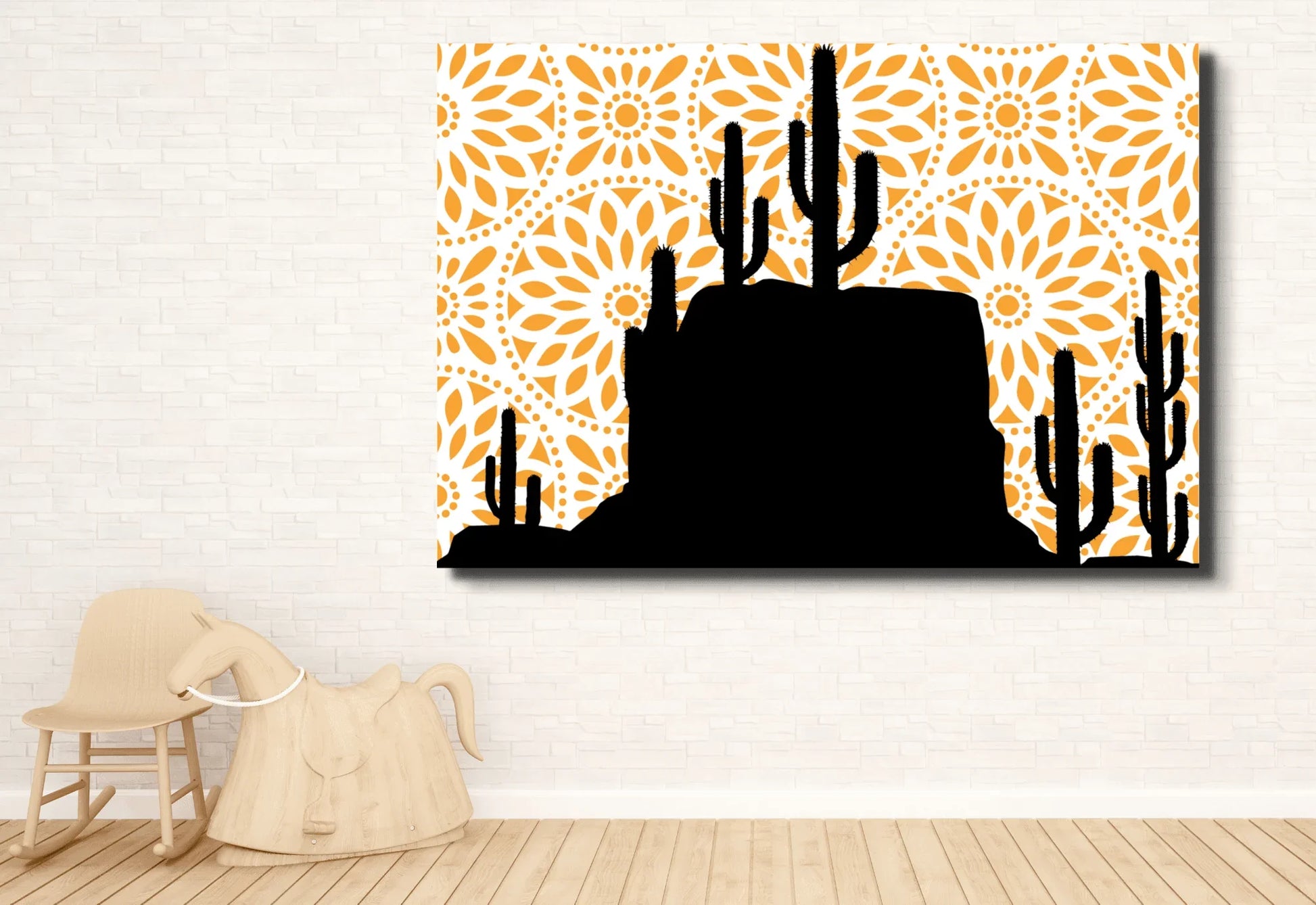 
A large canvas art print featuring a black silhouette of a desert landscape with saguaro cacti and a rocky mesa. The background is a bright orange mandala pattern with a white circular design. The print hangs on a white brick wall above a light wood rocking horse.
