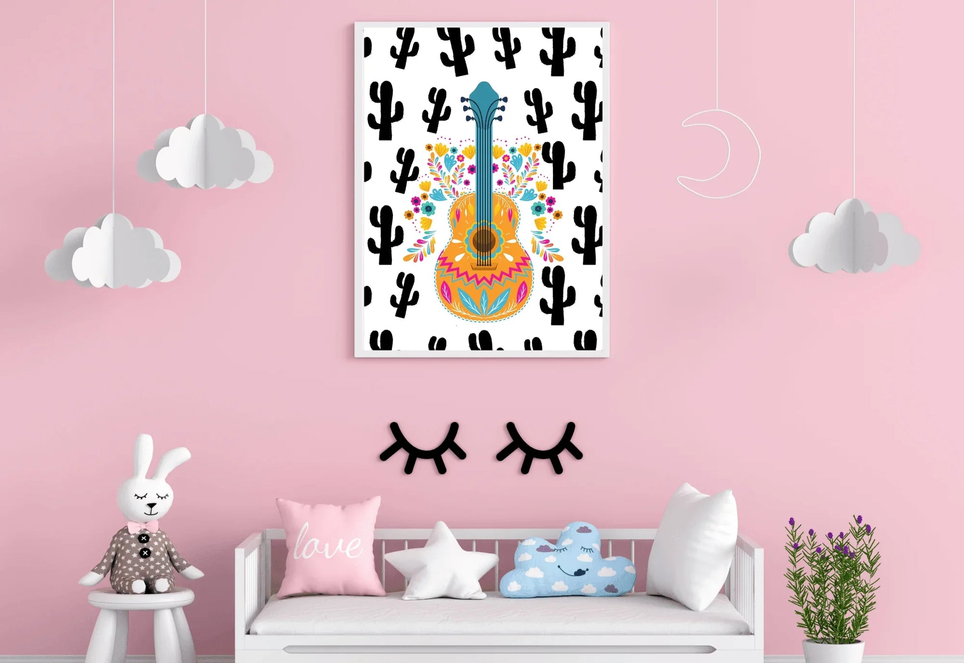 A framed boho folk art guitar print with a black cactus background, hanging on a light pink wall above a white bench with pillows.
