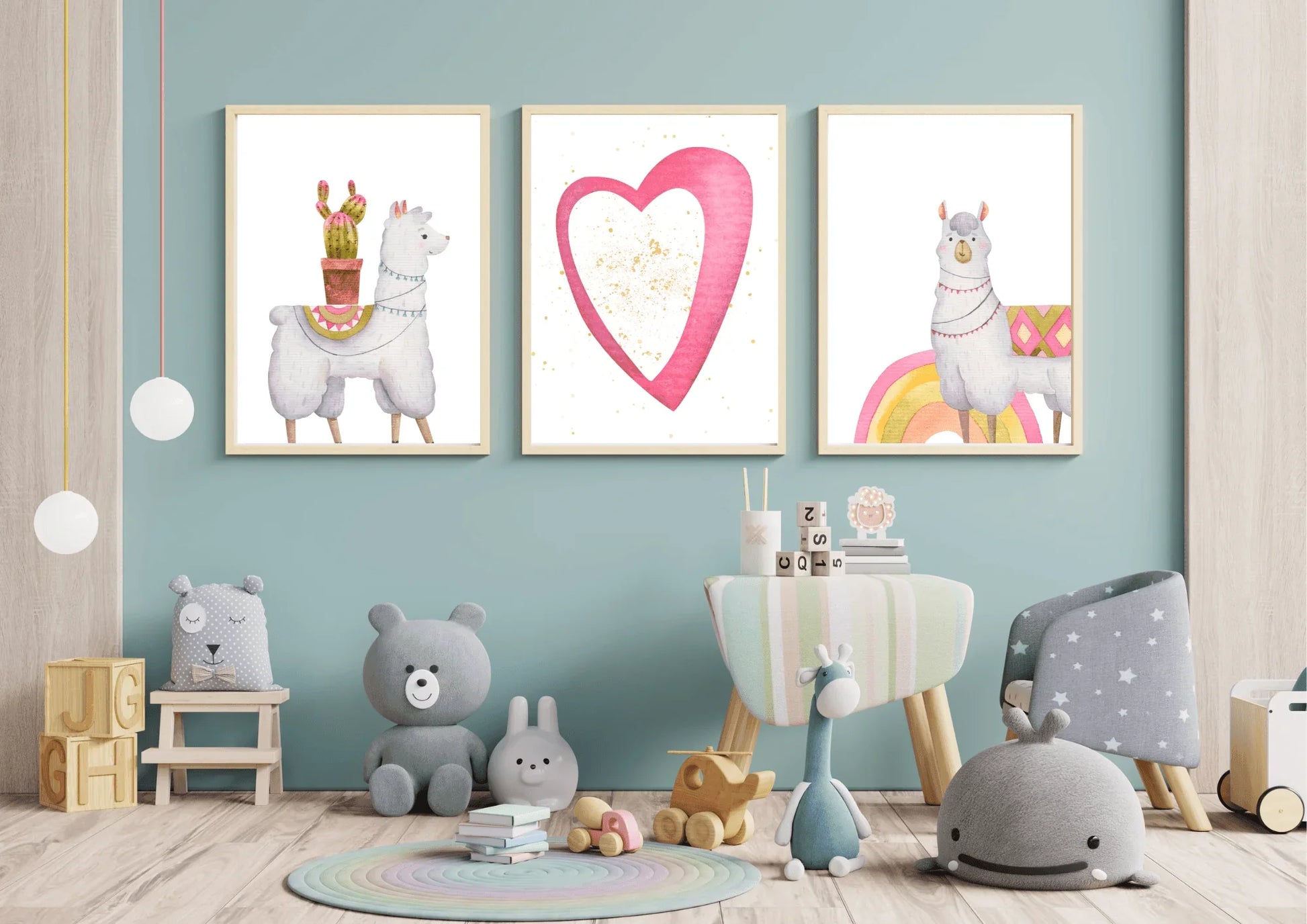 A trio of framed digital art prints hanging on a light blue wall in a child's room. The prints feature two llamas—one with a cactus on its back and another standing on a rainbow—and a central print of a large pink heart with gold speckles. The room is decorated with a round rug, a small chair and table, and various stuffed animals and toys.