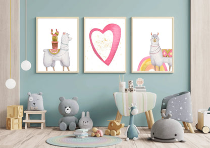 A trio of framed digital art prints hanging on a light blue wall in a child's room. The prints feature two llamas—one with a cactus on its back and another standing on a rainbow—and a central print of a large pink heart with gold speckles. The room is decorated with a round rug, a small chair and table, and various stuffed animals and toys.