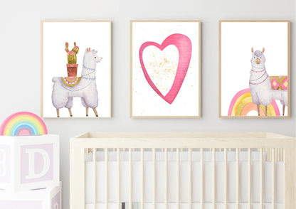 A trio of framed digital art prints hanging on a white wall above a baby crib. The prints feature two llamas with blankets—one carrying a cactus and another standing on a rainbow—and a central print of a pink heart with gold speckles. A set of large wooden blocks with letters is on the left.