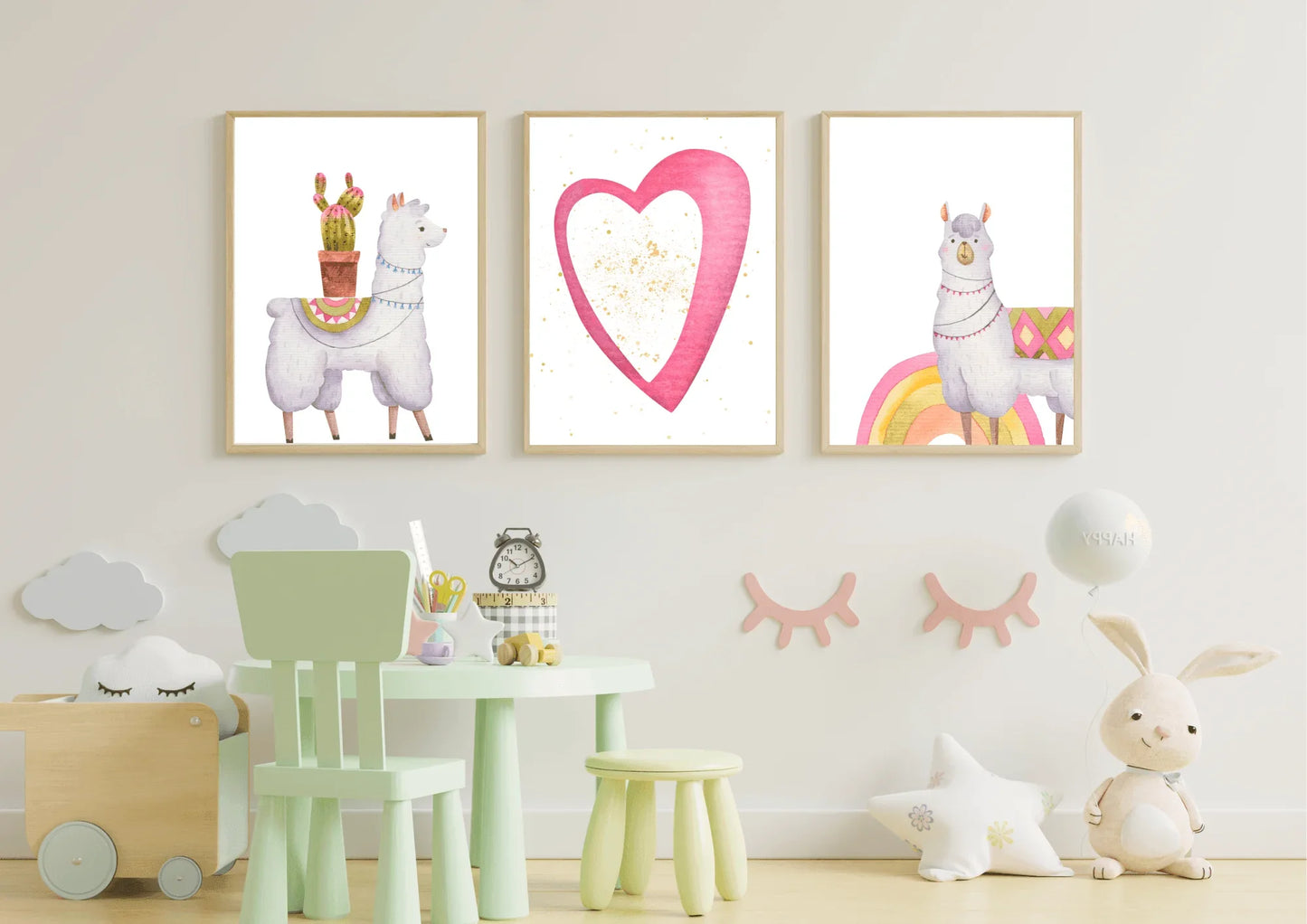 A set of three framed digital art prints hanging on a light-colored wall in a child's room. The prints feature two llamas and a large pink heart with gold speckles. A small green table with matching chairs, and a variety of toys and pillows are in the foreground.