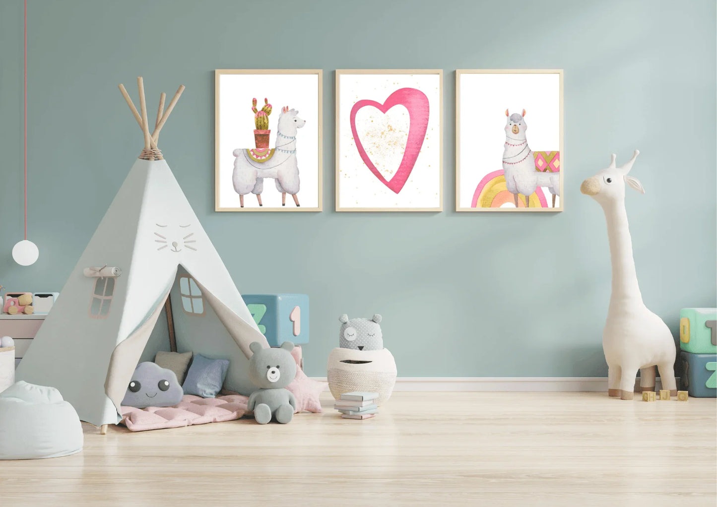 A set of three framed digital art prints featuring llamas and a heart, hanging on a light blue wall in a child's playroom. A white teepee tent, stuffed animals, and a large toy giraffe are arranged on the light wood floor.